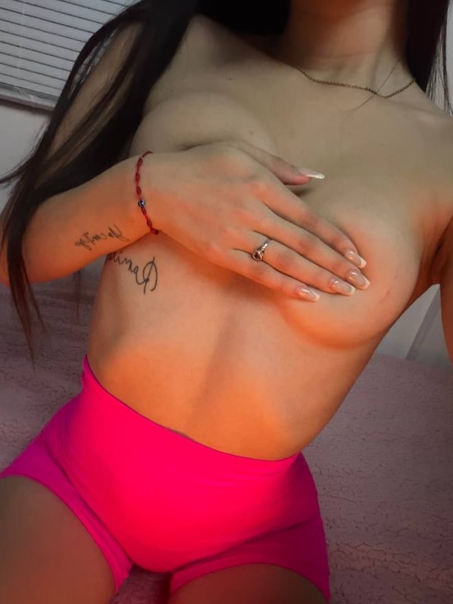 Young woman with long dark hair taking a mirror selfie in a bedroom, wearing a light pink tank top and white bottoms while kneeling on a fluffy pink rug.