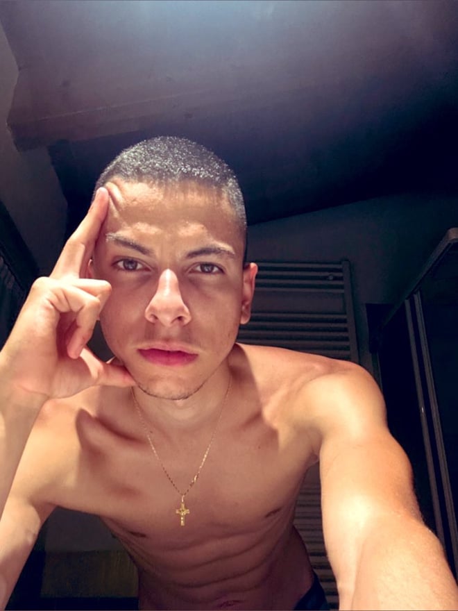 Young man with short hair posing shirtless indoors, hand on forehead, displaying muscular torso and necklace.