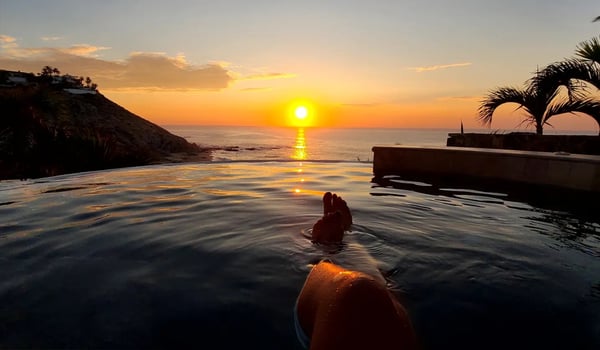 A person's legs and feet resting in an infinity pool overlooking a scenic sunset ocean view with palm trees.