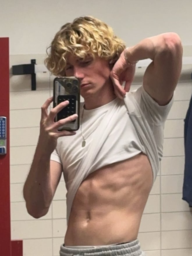 Young man with curly blonde hair taking a mirror selfie in a locker room, wearing a white t-shirt and grey sweatpants.