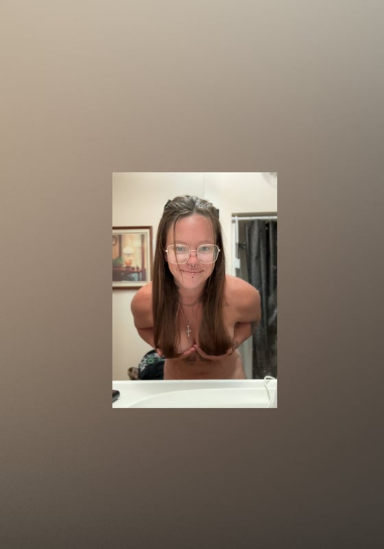 A woman with long hair and glasses smiling at the camera in an indoor setting with artwork on the wall.