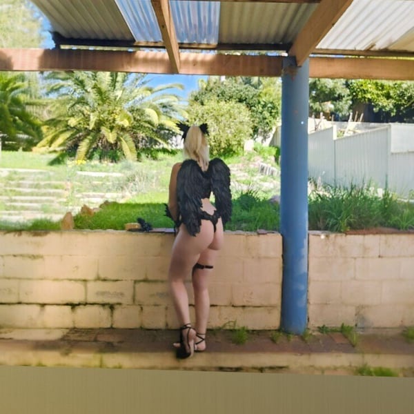 A woman dressed as a fallen angel with black wings and cat ears poses outdoors under a patio roof with palm trees and greenery in the background.