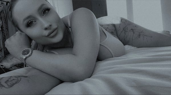 A woman with blonde hair reclining on a bed in a light-colored top, posing seductively with a watch on her wrist against a softly lit background.