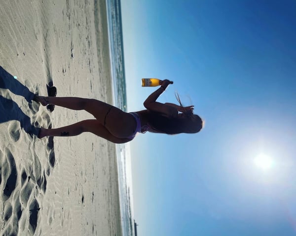 Young woman in a purple bikini posing on a sunny beach, holding a bottle and looking over her shoulder toward the ocean.