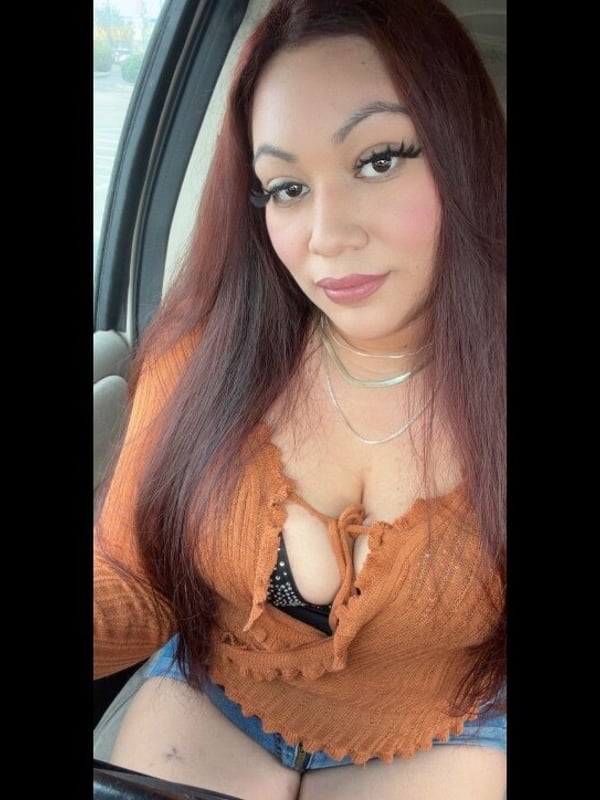 A woman with long dark hair sitting in a car, wearing an open orange cardigan and necklace, smiling at the camera.