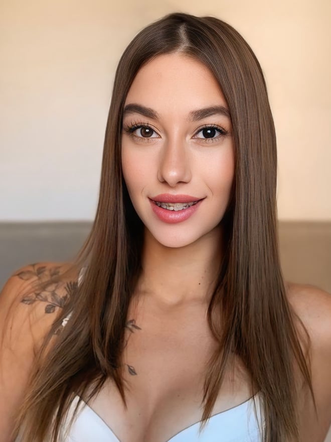 Close-up portrait of a smiling young woman with long brown hair and a shoulder tattoo, wearing a white lace top in a warmly lit room.
