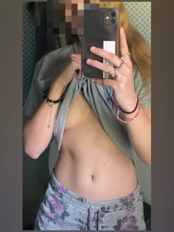 Young woman with blonde hair taking a mirror selfie in a bathroom, wearing an open gray shirt and patterned pants, with toned midriff visible.