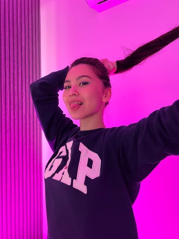 Young woman with long dark hair sticking out her tongue, wearing a navy GAP sweatshirt, posing against a pink background.