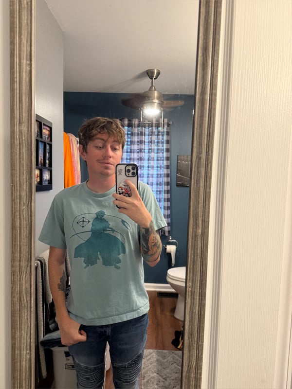 A person taking a selfie in a bathroom mirror, wearing a graphic t-shirt and jeans, with tattoos visible on the arm.