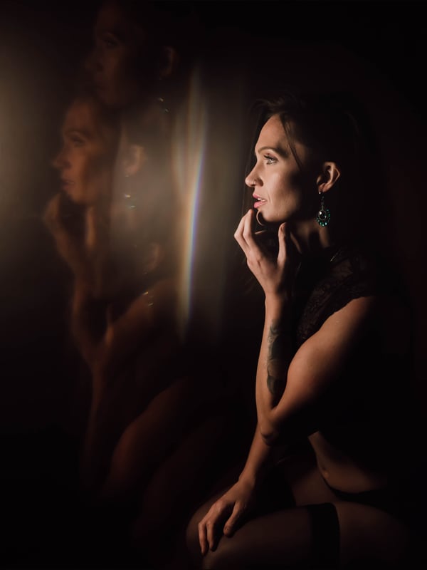 A woman seated in low light with a thoughtful expression, adorned with earrings and tattoos, in a shadowy indoor environment.