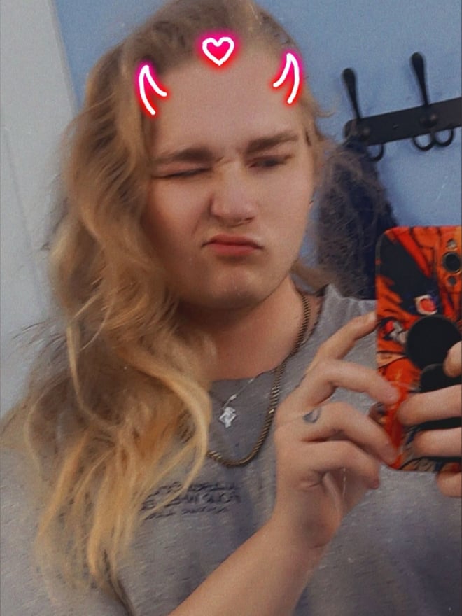A young man with long blonde hair wearing neon red devil horns filter, making a playful face while holding a phone against a bathroom background.