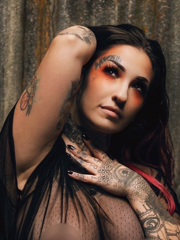 A woman with tattoos and bold makeup posing confidently against a rustic metal background, wearing a sheer black top.