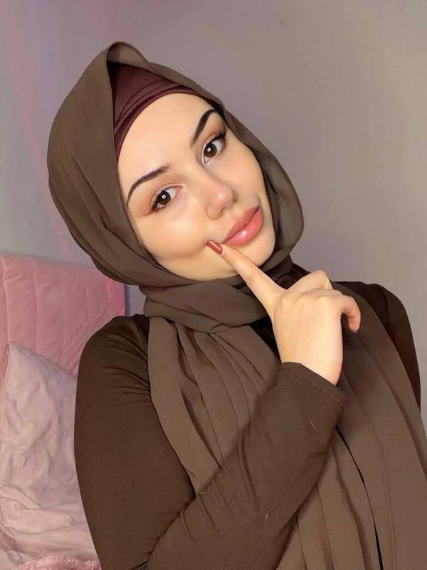 A woman wearing a brown headscarf and long-sleeved top, posing with a finger to her lips in a softly lit bedroom with pink bedding.