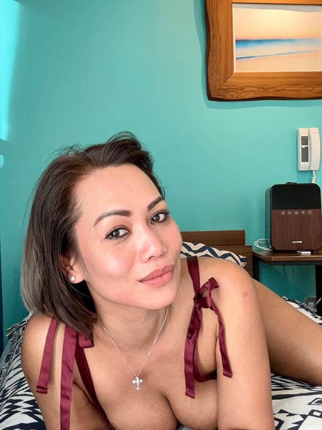 A woman reclining on a bed in a maroon ribbon top, smiling confidently in a cozy room with wooden decor and beach art.