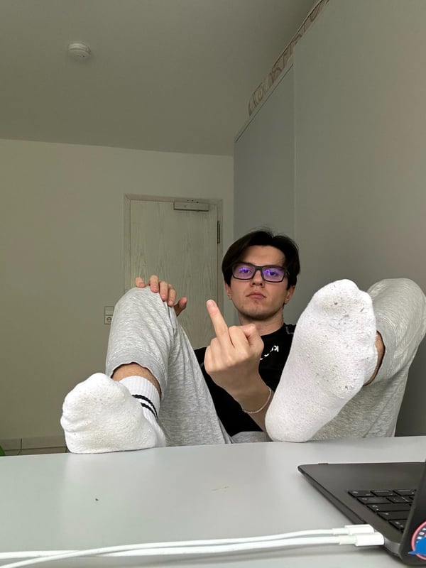 Young man sitting with legs raised, wearing glasses, a black shirt, gray sweatpants, and white socks, making a hand gesture in front of a laptop on a table.