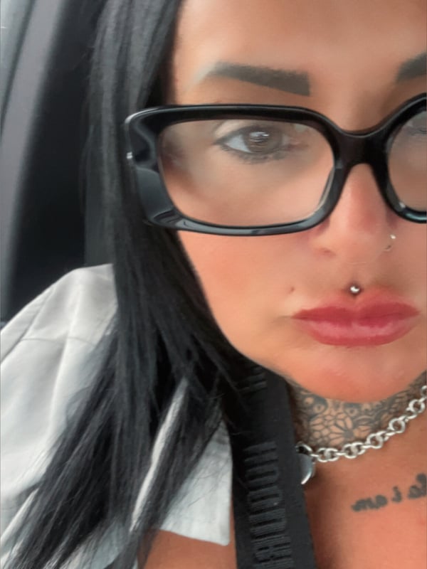 A woman with dark hair and glasses taking a close-up selfie in a car, wearing a choker necklace and showing neck tattoos.