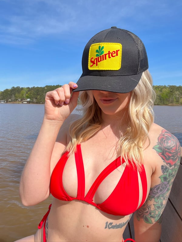 A woman with blonde hair wearing a red bikini top and a 'Squirt' trucker hat poses by a lake dock.