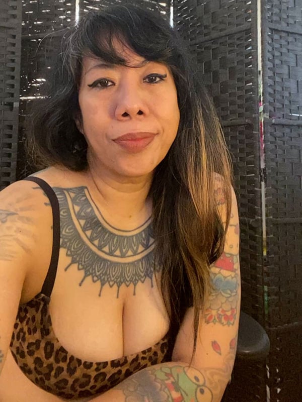 Woman with dark hair and visible tattoos posing in front of a bamboo screen, wearing a leopard print top and necklace.