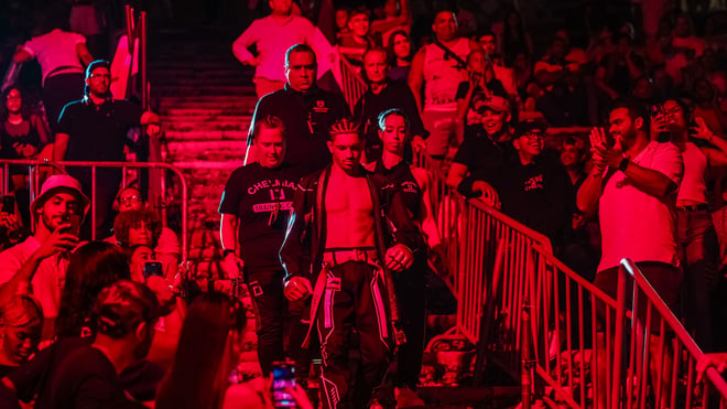 A muscular fighter walking shirtless through a crowd in a brightly lit arena during an event entrance.