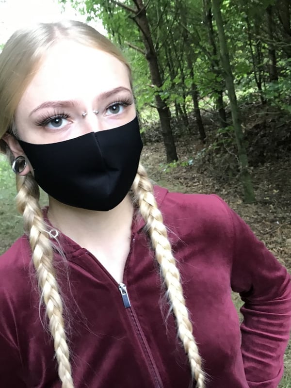 Young woman with blonde pigtails and glasses, wearing a black face mask and maroon velour tracksuit top, standing in a forested area with trees in the background.