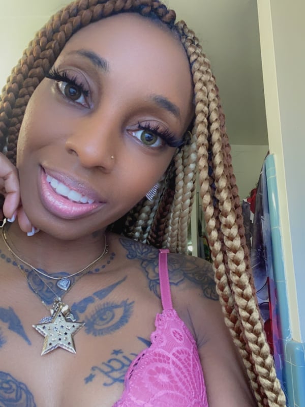 A close-up of a woman with braided hair and pink lipstick smiling at the camera, wearing a lace top and jewelry.