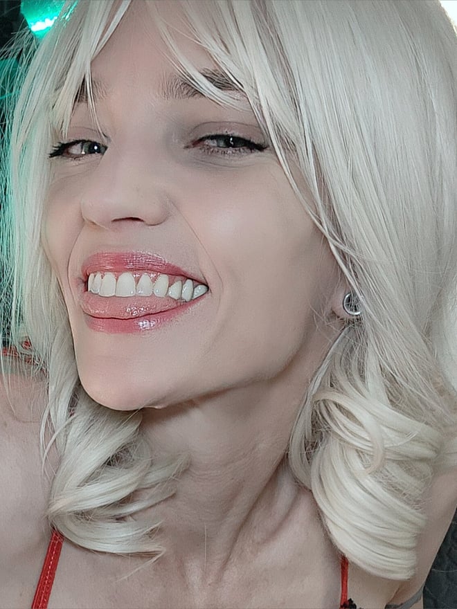 A close-up portrait of a woman with long blonde wavy hair, smiling with tongue out, illuminated by green neon lights.