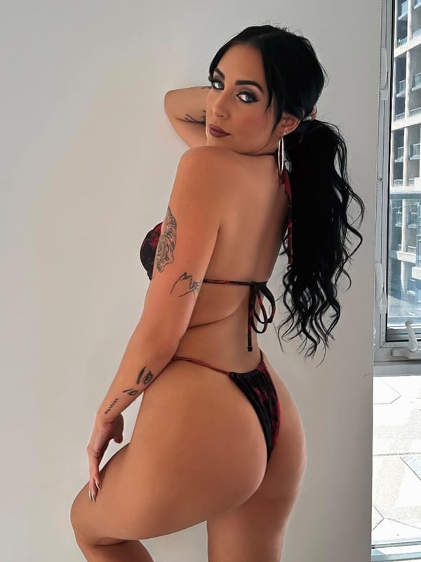 Woman standing sideways in a red bikini, highlighting her figure and tattoos indoors near a window.