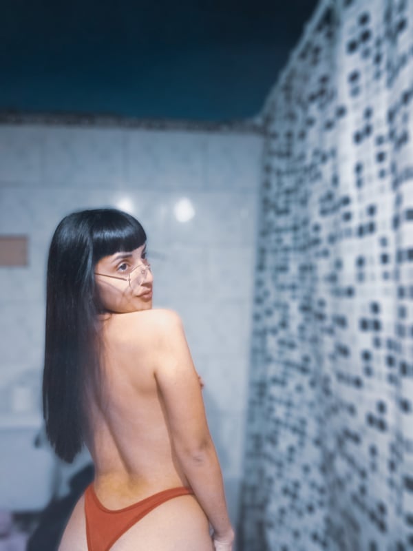 A woman with long dark hair stands with her back turned in a bathroom, wearing a red swimsuit bottom against a tiled wall.