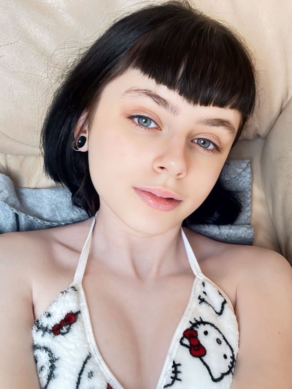Close-up of a young woman with dark bob hair lying on a bed, wearing a white top with red heart patterns, smiling gently.