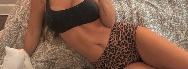 A woman lounging on a bed in a black tank top and leopard print bottoms, posing confidently with one hand on her hip.
