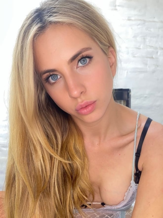 A blonde woman with blue eyes poses in a close-up portrait while wearing light-colored clothing.