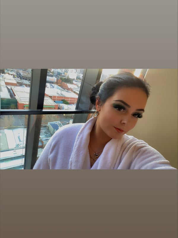 A woman with makeup wearing a white robe, smiling at the camera near a window.