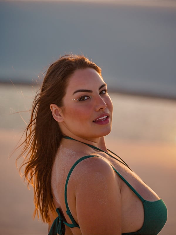 Close-up portrait of a smiling woman with long brown hair wearing a green bikini top against a beach sunset.
