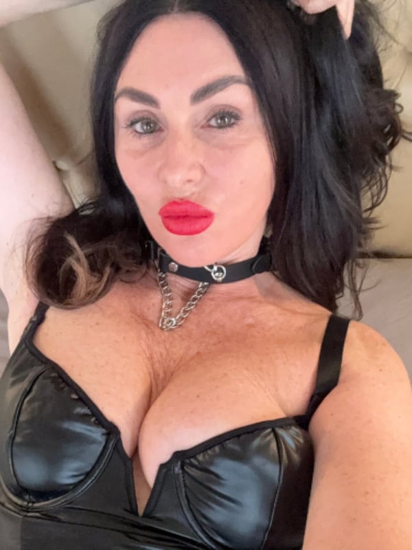 A woman with long dark hair posing confidently on a bed in elegant black clothing and a choker necklace.