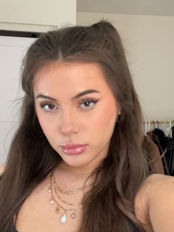 Young woman with long brown hair in pigtails, wearing layered necklaces, posing in front of a clothing rack indoors.