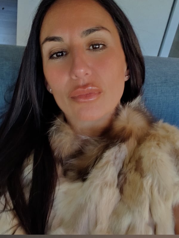 A woman with long dark hair wearing a fur-trimmed top, seated on a blue couch with a serious expression.