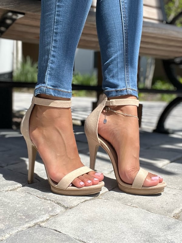 Close-up of a woman's legs and feet in nude high heels and jeans against an outdoor bench.