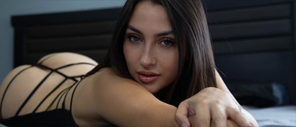 A woman lying on a bed in lingerie, posing confidently with a sultry expression against a dark bedroom background.