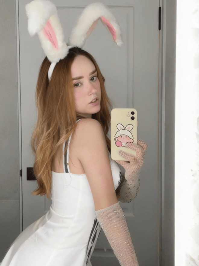 A young woman with long auburn hair takes a mirror selfie while wearing a white bunny-ear headband and white outfit.
