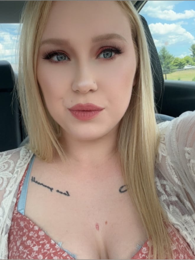 A woman with blonde hair taking a selfie in a car, wearing a lace cover-up over a top, with tattoos visible on her arms.