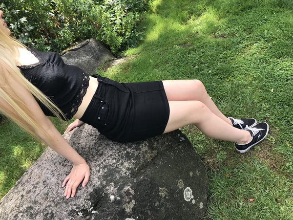 Young woman with long blonde hair lying on a large rock outdoors, wearing a black lace top and short skirt with sneakers, surrounded by green grass and bushes.