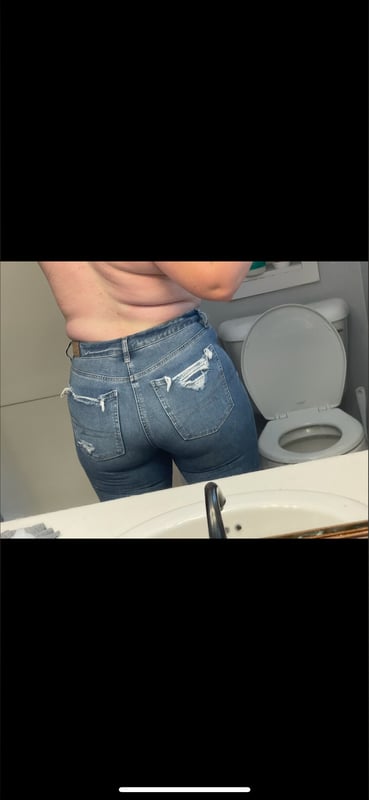 A woman standing in a bathroom, wearing ripped jeans, viewed from behind near a sink and toilet.