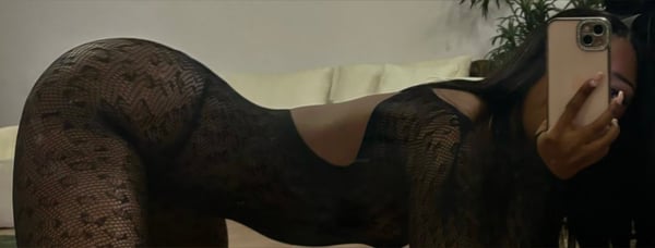 A person posing on a couch in a lacy black outfit, holding a phone for a selfie with plants in the background.