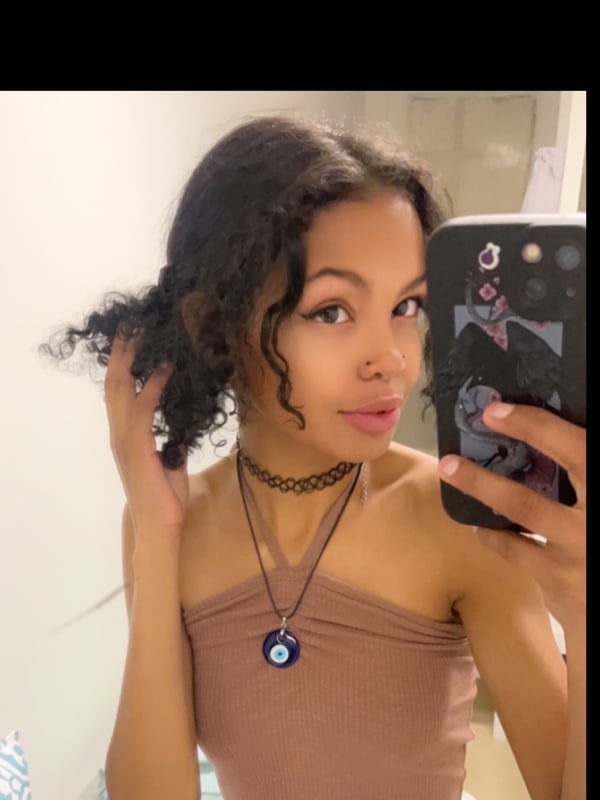 Young woman with curly dark hair taking a mirror selfie, wearing a beige tube top and choker necklace while holding her phone.