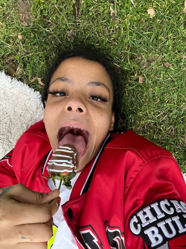 Young woman lying on grass outdoors, wearing a red Chicago Bulls jacket, holding and eating a chocolate-covered strawberry on a stick.