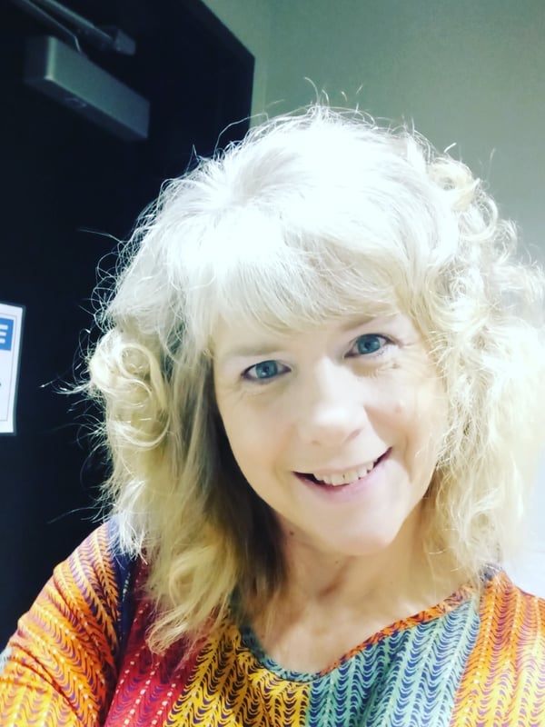 A smiling woman with curly blonde hair wearing a colorful striped top, standing indoors near a door with a notice sign and blue decorative item.