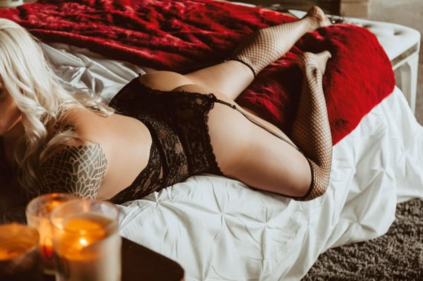 A woman with blonde hair reclining on a bed with red blankets and fishnet stockings, surrounded by candles in a cozy bedroom setting.