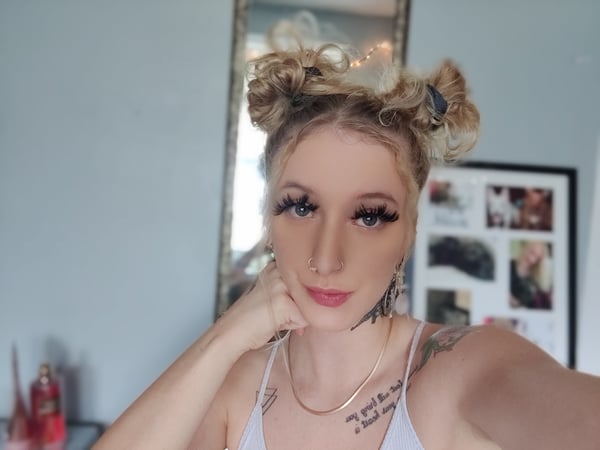 A young woman with blonde hair in space buns poses thoughtfully in a mirror selfie, wearing a light-colored top in a cozy indoor room with framed photos.