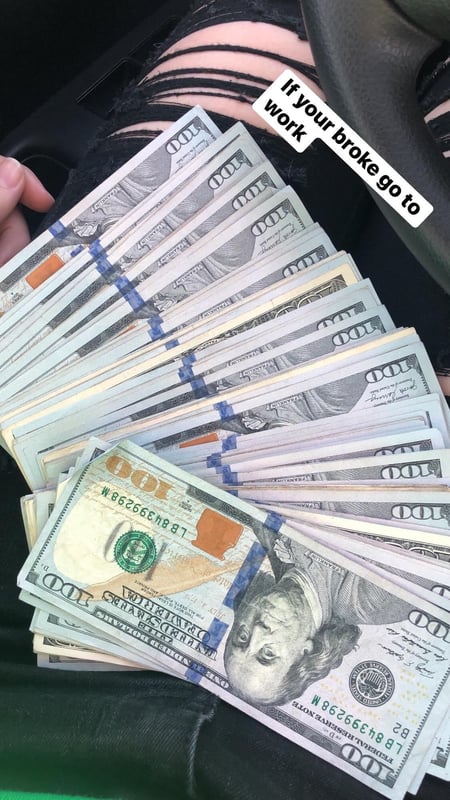 A person sitting in a car holding a stack of cash with a green background visible.