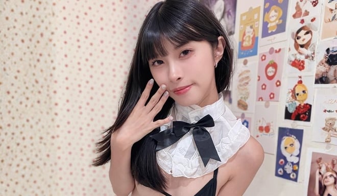 A young woman with long dark hair poses in a decorative room, wearing a white ruffled top with a black bow.